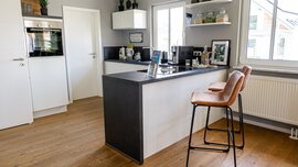 Moderne Küche mit Holzboden, weißen Schränken, schwarzer Arbeitsplatte, Barhockern und Dekorationsgegenständen auf Regalen.
