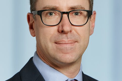 Mann mit kurzen dunklen Haaren, schwarz gerahmter Brille und Anzug mit Krawatte posiert lächelnd.
