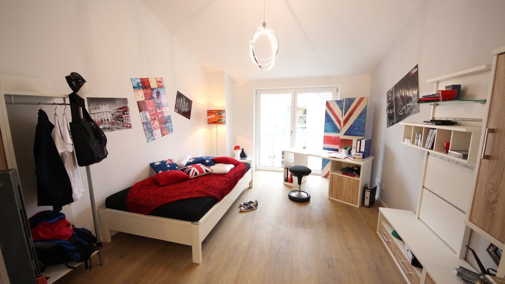 Blick in ein eingerichtetes Jugendzimmer mit Bett, Schreibtisch, Fernsehwand, Kleiderstange und bodentiefem Fenster.