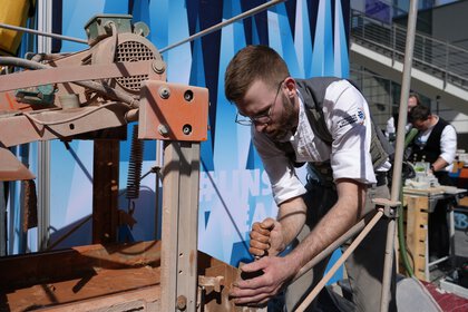 Ein Mann mit Brille und in Kluft bedient eine braune Maschine an einem Stand auf der BAU.