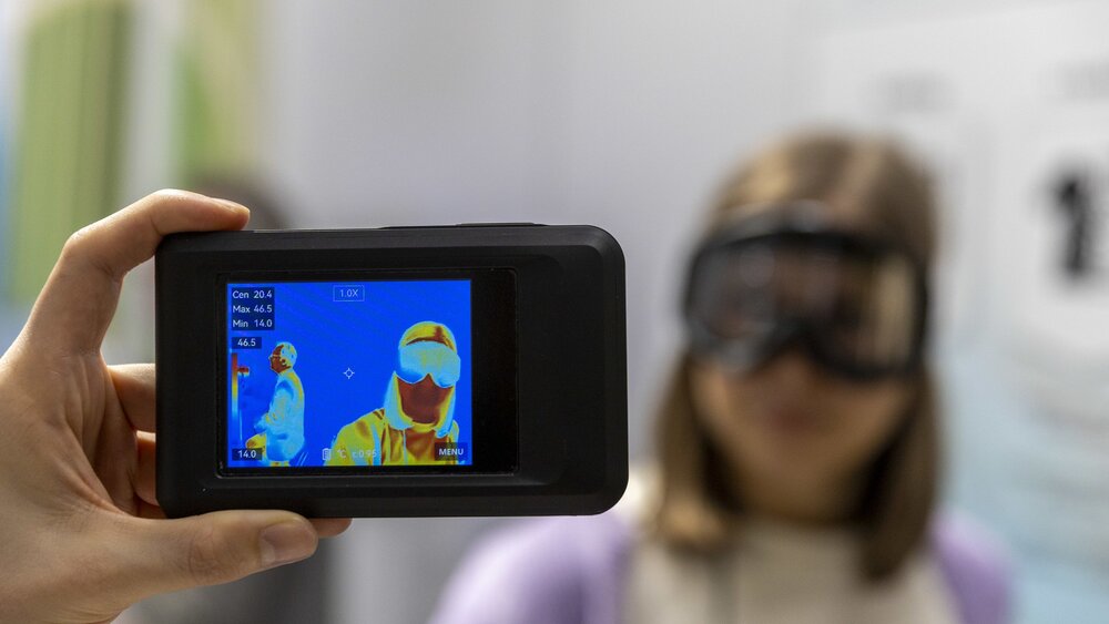 Eine Hand hält eine Wärmebildkamera mit Vorschaudisplay und richtet sie auf eine Frau mit einer Skibrille.