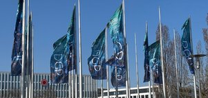 Mehrere Fahnenmasten mit Fahnen, auf denen die Initialen „DGP“ zu lesen sind, wehen vor weißen Gebäuden und blauem Himmel.