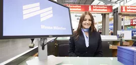 A woman in a suit and scarf sits at an information desk with a monitor displaying the Messe München logo.