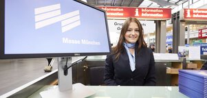 Eine lächelnde Frau mit langen braunen Haaren, einem Anzug und einem blauen Halstuch sitzt an einem Informationsschalter der Messe München, neben einem Display.