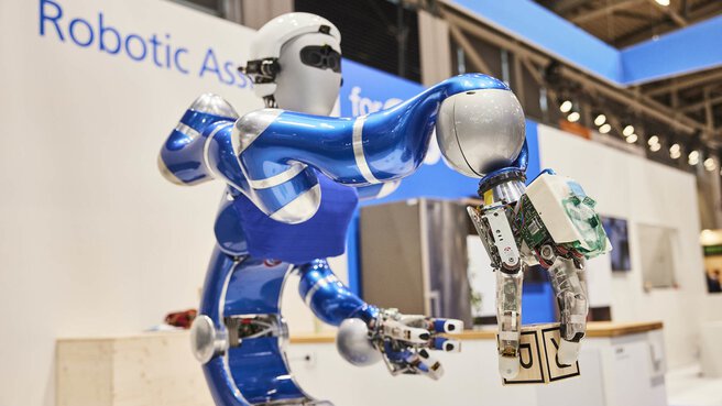 Robot with a humanoid upper body on display at a trade fair stand, holding a blue block in its gripper.
