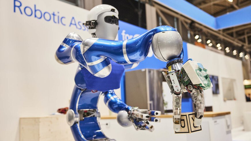 Roboter mit humanoidem Oberkörper, der an einem Messestand ausgestellt ist und einen Blauklotz im Greifer hält.