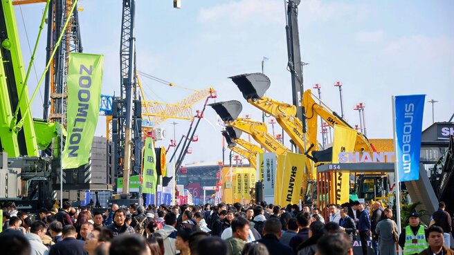 On a sunny day, a large crowd strolls among cranes and construction equipment at an outdoor fair.