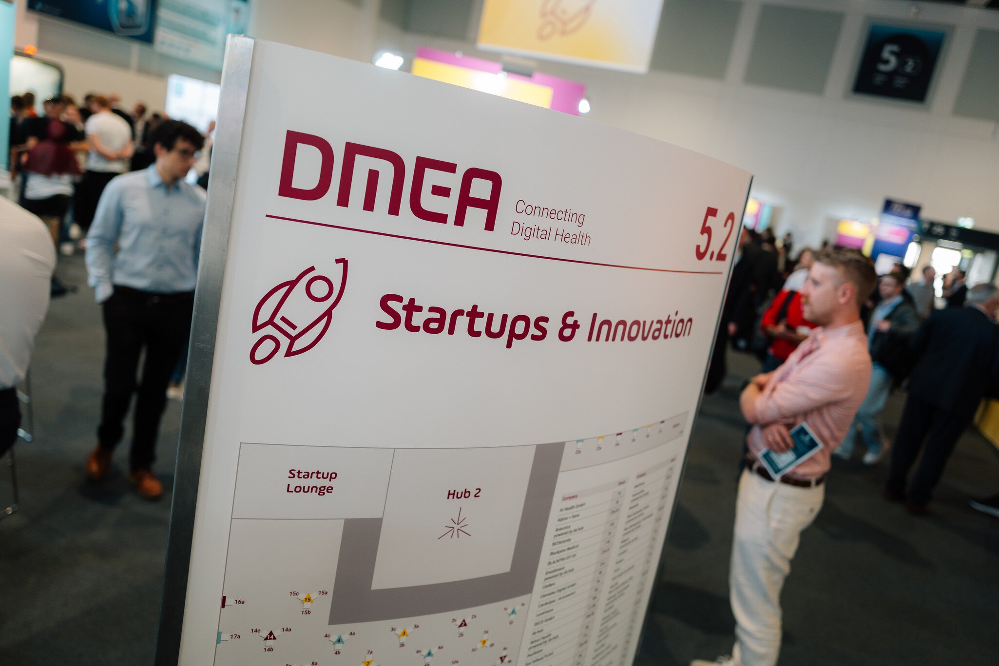 DMEA Schild für den Bereich Startups & Innovation mit Hallenplan und Besuchern im Hintergrund