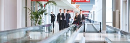 A group of people in business attire is walking along the express walkways between the exhibition halls at Messe München. Red directional signs hang above them.