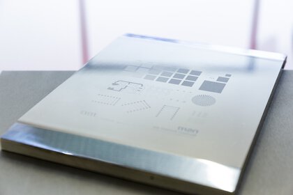 Silbernes Metallblech mit eingravierten geometrischen Mustern und Formen auf der Oberfläche, auf einem grauen Tisch liegend.