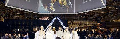 Several models, dressed in light-coloured clothing and wearing jewellery, are standing together in a circle, under several video screens, facing an interested audience.