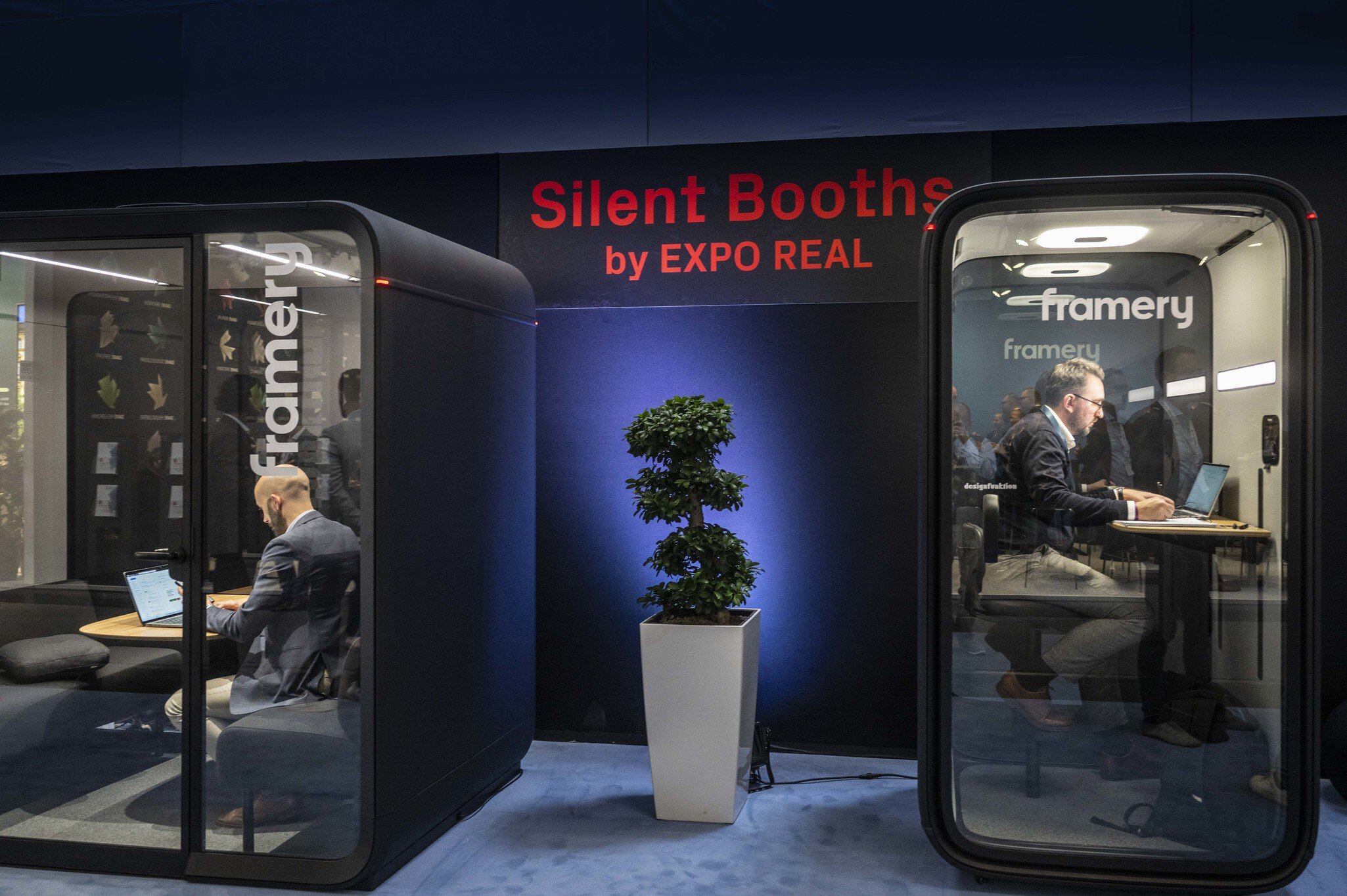 Two men are working inside soundproof booths (“Silent Booths by EXPO REAL”) at EXPO REAL in Munich. Both are seated and focused on their laptops in modern glass work pods by the brand “framery.” A green plant stands between the booths, and the scene is lit with blue light, creating a calm and productive atmosphere.