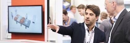 Zwei Männer im Anzug im Gespräch auf einer Photonik-Messe, einer erklärt ein Gerät am Stand, im Hintergrund Aussteller, Display und Messeschilder.