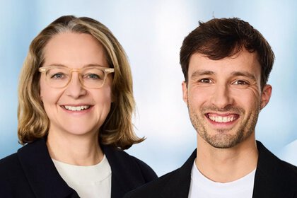 Portrait of two smiling people against a blue-white background, a woman with blond hair and glasses, and a man with dark hair and short beard.