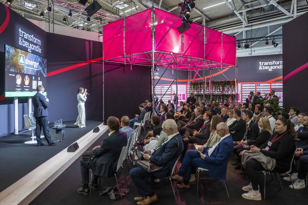 A woman speaks on stage to a seated audience in a modern conference hall with light pink décor.