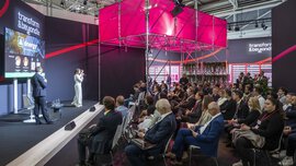 A woman speaks on stage to a seated audience in a modern conference hall with light pink décor.