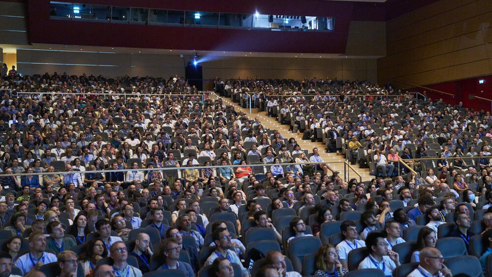 A large audience sits in an auditorium and attentively follows a presentation.