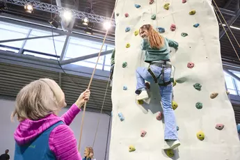 Eine Person klettert an einer Indoor-Kletterwand hoch und schaut nach unten zu der Person, von der sie gesichert wird.