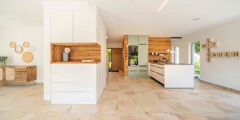 Ein offener Wohnraum mit Steinfußboden, einem breiten Küchentresen, weißen Schränken, Holzvertäfelung und bodentiefen Fenstern.