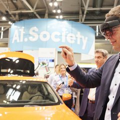 A man in a suit wears augmented reality glasses next to an orange car at a trade fair stand and points to the air in front of him with one hand.