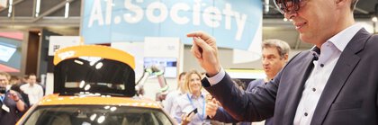 A man in a suit wears augmented reality glasses next to an orange car at a trade fair stand and points to the air in front of him with one hand.
