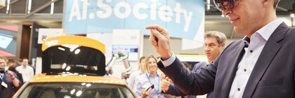 A man in a suit wears augmented reality glasses next to an orange car at a trade fair stand and points to the air in front of him with one hand.