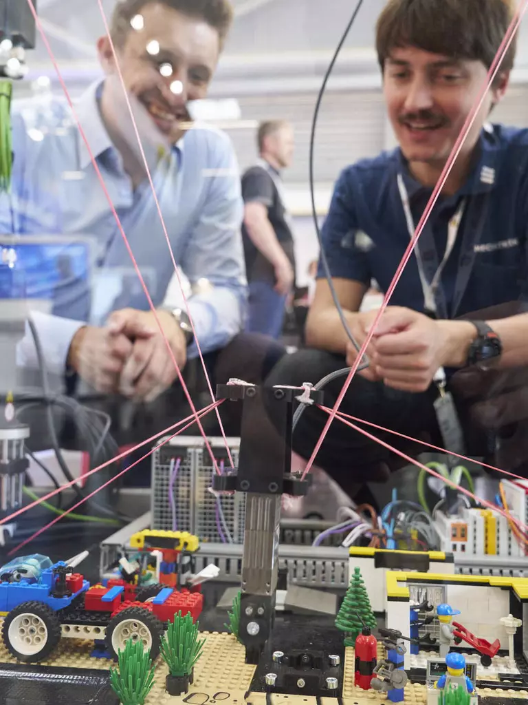 Zwei Männer betrachten einen in einer Vitrine aufgebauten Kabelroboter, der mit Legosteinen interagiert.