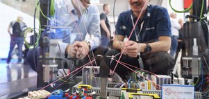 Zwei Männer betrachten einen in einer Vitrine aufgebauten Kabelroboter, der mit Legosteinen interagiert.