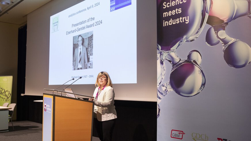 a person stands behind a lectern; in the background you can see a presentation