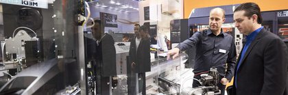 Two men stand in front of a machine exhibited in a reflective display case at a productronica trade fair stand. The man on the left points to a detail.