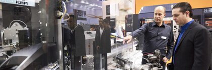 Zwei Männer stehen vor einer Maschine, die an einem Messestand der productronica in einer spiegelnden Vitrine ausgestellt ist. Der linke Mann deutet auf ein Detail.