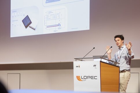 Ein Mann an einem Rednerpult hält eine Präsentation vor einer großen Leinwand. Das Publikum befindet sich außerhalb des Bildes.