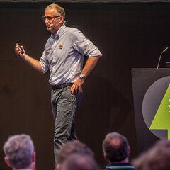 A man with glasses speaking in front of an audience and gesticulating with his hand. On the right is a podium with the INTERFORST logo.