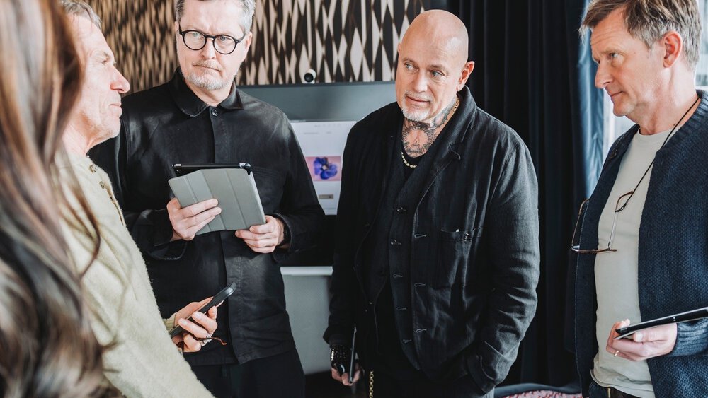 Five people are standing together around a low wooden table with several pieces of jewelry in black wooden boxes and are talking to each other.