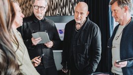 Five people are standing together around a low wooden table with several pieces of jewelry in black wooden boxes and are talking to each other.
