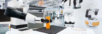Zwei Roboter in weiß und schwarz mit entfernt humanoiden Zügen stehen in einer Werkshalle. Der eine Roboter greift mit seinem Arm nach einem orangenen Zylinder.