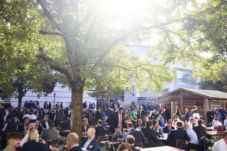 Outdoor area of Messe München, where numerous visitors sit at catering tables under the trees and chat.