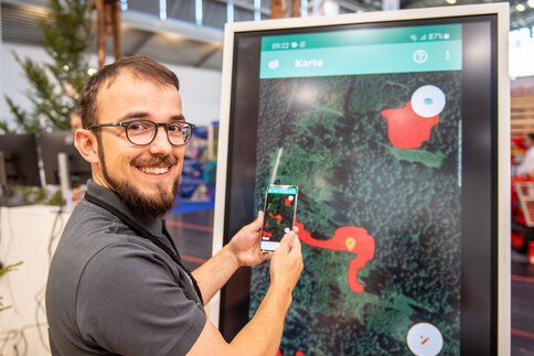  Mann bedient ein interaktives Display mit digitaler Karte und hält ein Smartphone in der Hand, im Hintergrund Messestand