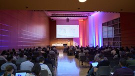 Atmospheric, colorfully lit conference room with seated participants watching a speaker on stage and a projection screen.