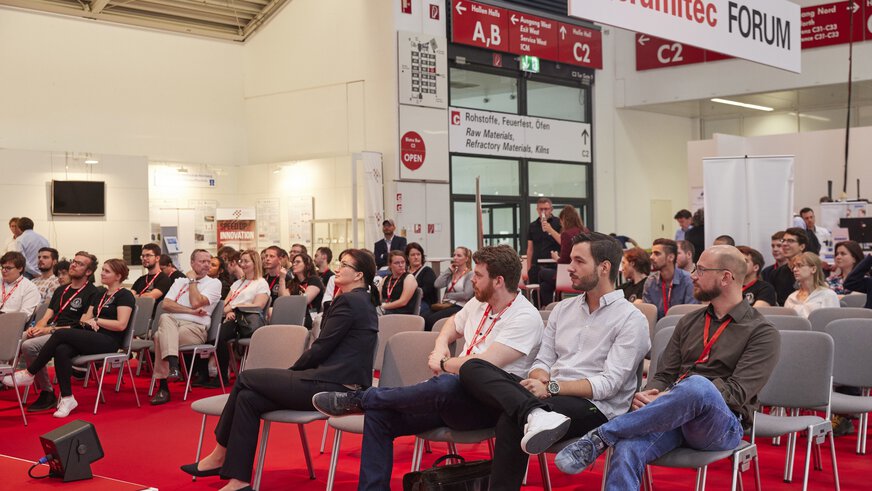 A large audience sits in rows at a lecture in the exhibition hall. Some people are holding notepads in their hands.