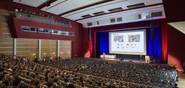 Zahlreiche Sitzreihen mit Publikum vor einer Bühne und einer großen Leinwand, auf der eine Präsentation läuft.