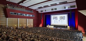 Numerous rows of seats with an audience in front of a stage and a large screen showing a presentation.