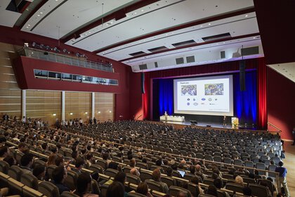 Zahlreiche Sitzreihen mit Publikum vor einer Bühne und einer großen Leinwand, auf der eine Präsentation läuft.