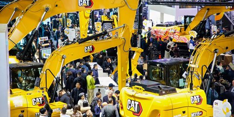 Crowds gather around large yellow CAT excavators at a bustling construction equipment trade show.