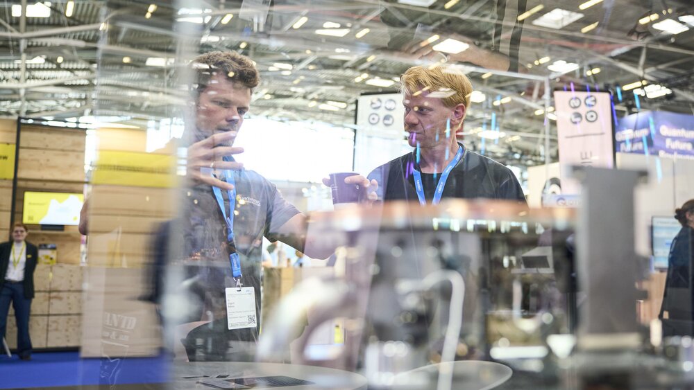 Zwei Männer diskutieren angeregt hinter einer Glasvitrine mit Technikexponat auf der World of Quantum 2025 in München.