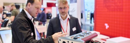 Zwei Männer im Anzug diskutieren über ein mechanisches Bauteil, das auf der Messe ausgestellt ist.