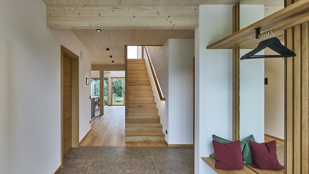 Moderner Flur mit Holztreppe, Bank mit bunten Kissen und großen Fenstern, die in einen offenen, hellen Raum führen.