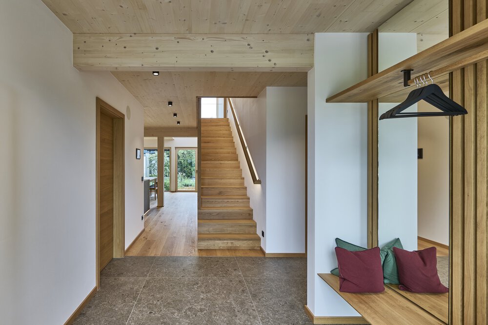 Moderner Flur mit Holztreppe, Bank mit bunten Kissen und großen Fenstern, die in einen offenen, hellen Raum führen.