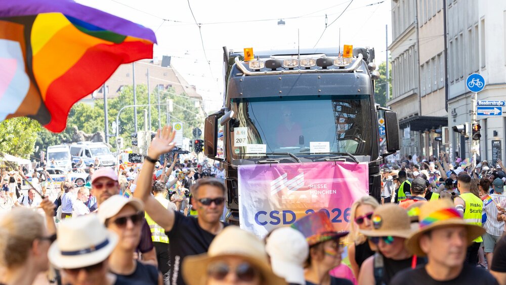 During a Pride parade, people drive through the city in a truck decorated with rainbow flags and celebrate on a sunny street.
