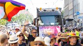 Die Menschen ziehen bei einer Pride-Parade mit einem Lastwagen und Regenbogenfahnen durch die Stadt und feiern auf einer sonnigen Straße.